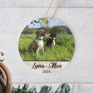May include: A round ceramic ornament featuring a photograph of a tan and white dog and a black and white dog sitting in a grassy field. The bottom section displays the text Luna - Max and the year 2023 in black font on a light wood background.