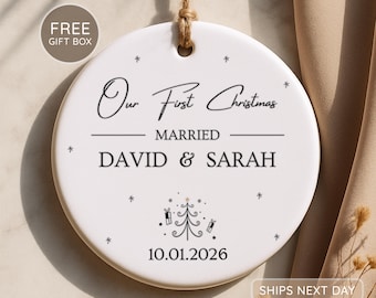 May include: A round white ceramic ornament hanging from a jute twine loop. The text reads Our First Christmas MARRIED DAVID & SARAH with the date 10.01.2026. A small black line drawing of a Christmas tree with gifts and star accents is centered.