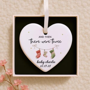 Op de afbeelding: Een wit hartvormig ornament met de tekst "AND THEN there were three" en "baby charlie 12.17.25". Het ornament heeft drie hangende kousen in rood, wit en groen. Gepresenteerd in een geschenkdoos met een zwart interieur en een wit lint.
