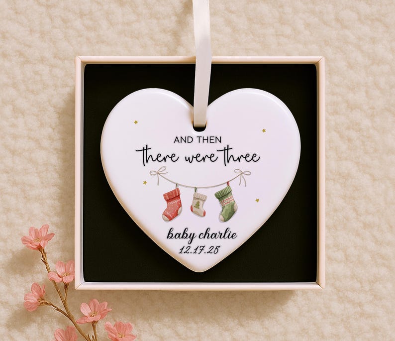 Op de afbeelding: Een wit hartvormig ornament met de tekst "AND THEN there were three" en "baby charlie 12.17.25". Het ornament heeft drie hangende kousen in rood, wit en groen. Gepresenteerd in een geschenkdoos met een zwart interieur en een wit lint.