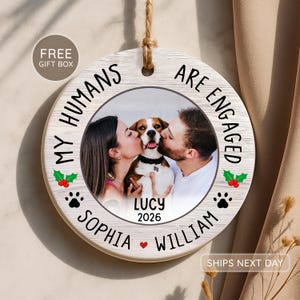 Op de afbeelding: Een rond ornament met houtstructuur hangend aan een jute touwtje. Het midden toont een foto van een koppel dat een hond kust. De tekst luidt: MY HUMANS ARE ENGAGED, SOPHIA, WILLIAM, LUCY, 2026. Versierd met hulstblaadjes, rode bessen en zwarte pootafdrukken.