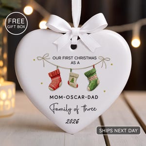 K&ouml;nnte beinhalten: Wei&szlig;er herzf&ouml;rmiger Weihnachtsschmuck mit wei&szlig;er Schleife. Der Schmuck tr&auml;gt den Text "OUR FIRST CHRISTMAS AS A" und "Family of three 2026". Drei h&auml;ngende Str&uuml;mpfe sind ebenfalls abgebildet.