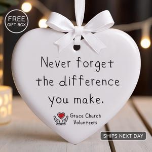 May include: A white heart-shaped ornament with a white ribbon and bow. The ornament has the text "Never forget the difference you make." and the Grace Church Volunteers logo. A "FREE GIFT BOX" label is in the upper left corner.