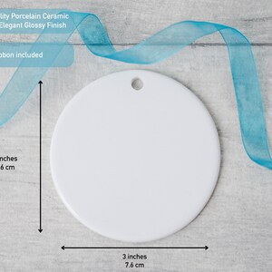 May include: A round white porcelain ceramic ornament with a glossy finish and a small hole at the top for hanging. The ornament measures 3 inches in height and 3 inches in width. A blue ribbon is included with the ornament for display purposes.