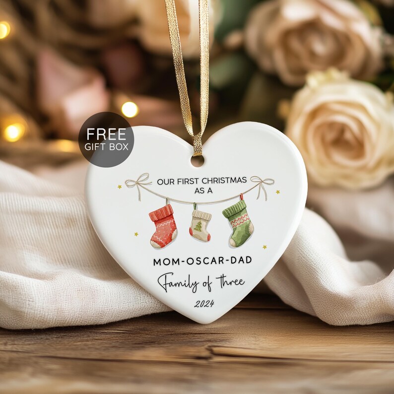 K&ouml;nnte beinhalten: Weihnachtsornament in Herzform mit goldenem Band. Das wei&szlig;e Keramikornament tr&auml;gt den Text "OUR FIRST CHRISTMAS AS A" und "MOM-OSCAR-DAD Family of three 2024". Drei Str&uuml;mpfe sind in Rot, Wei&szlig; und Gr&uuml;n dargestellt.