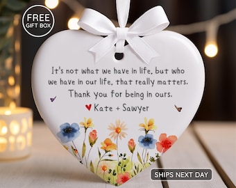 Op de afbeelding: Wit hartvormig ornament met een bloemenontwerp en een wit lint. Het ornament heeft de tekst "Het is niet wat we in het leven hebben, maar wie we in ons leven hebben, dat er echt toe doet. Bedankt dat je in de onze bent. Kate + Sawyer."