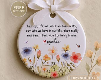 Op de afbeelding: Ronde, witte keramische ornament met een bloemenontwerp en een wit lint om op te hangen. Het ornament heeft de tekst: "Ashley, het is niet wat we in het leven hebben, maar wie we in ons leven hebben, dat er echt toe doet. Bedankt dat je van mij bent. Sophia."