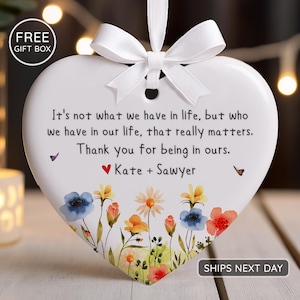 May include: White heart-shaped ornament with a floral design and a white ribbon. The ornament has the text "It's not what we have in life, but who we have in our life, that really matters. Thank you for being in ours. Kate + Sawyer."