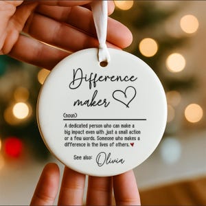 May include: A white, round ceramic ornament with the words "Difference maker" in black script, along with a heart symbol. The ornament includes a definition and the name "Olivia". A white ribbon is attached for hanging.
