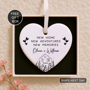 May include: A white heart-shaped ornament with a ribbon for hanging, presented in a gift box. The ornament features the text "NEW HOME NEW ADVENTURES NEW MEMORIES" and the names "Olivia + William" with a house graphic and the year 2026.