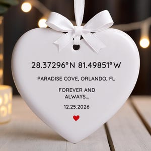 May include: A white heart-shaped ornament with a white ribbon bow at the top. The ornament has text that includes coordinates, location, and the phrase "FOREVER AND ALWAYS..." with a date and a small red heart.