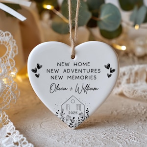 May include: White heart-shaped ceramic ornament with a jute string for hanging. The ornament features black text that reads "NEW HOME, NEW ADVENTURES, NEW MEMORIES" and the names "Olivia + William" with a house graphic and the year 2025.