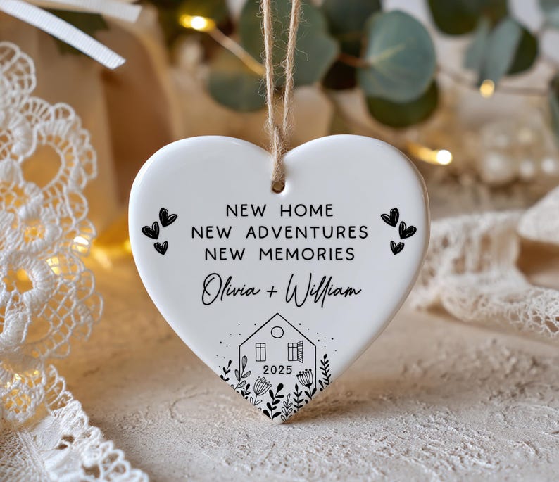 May include: White heart-shaped ceramic ornament with a jute string for hanging. The ornament features black text that reads "NEW HOME, NEW ADVENTURES, NEW MEMORIES" and the names "Olivia + William" with a house graphic and the year 2025.