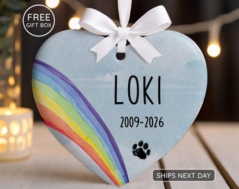Könnte beinhalten: Ein herzförmiges Keramikornament mit einem Aquarell-Regenbogen auf der linken Seite vor einem hellblauen Hintergrund. Der Text LOKI und 2009-2026 ist in Schwarz aufgedruckt, ergänzt durch einen kleinen schwarzen Pfotenabdruck. Ein weißes Band ist oben zu einer Schleife gebunden.