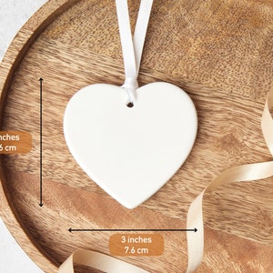 May include: A white heart-shaped ornament, 3 inches (7.6 cm) wide, with a white ribbon for hanging. The ornament is displayed on a wooden tray with a cream-colored ribbon.