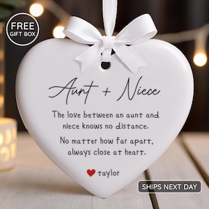 May include: White heart-shaped ornament with a white ribbon and bow. The ornament has the words "Aunt + Niece" and a message about love. A red heart and the name "taylor" are also on the ornament.