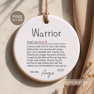 May include: A round white ceramic ornament hanging from a jute twine loop. The text reads Warrior, followed by a phonetic pronunciation with a red heart, a definition of a warrior, the text See also: Angie, and the year 2026 in black ink.