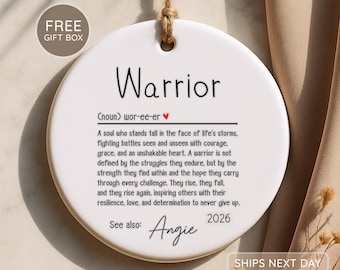 Peut inclure: Une décoration ronde en céramique blanche suspendue par une boucle en corde de jute. Le texte indique Warrior, suivi d'une prononciation phonétique avec un cœur rouge, une définition, See also: Angie et l'année 2026 en encre noire.