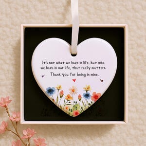 May include: A white heart-shaped ornament with a ribbon for hanging, presented in a box. The ornament features a floral design at the bottom and a heartfelt message: "It's not what we have in life, but who we have in our life, that really matters. Thank you for being in mine."