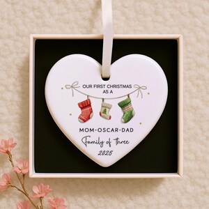 K&ouml;nnte beinhalten: Ein wei&szlig;er, herzf&ouml;rmiger Weihnachtsschmuck mit einem Band zum Aufh&auml;ngen. Der Schmuck zeigt den Text "OUR FIRST CHRISTMAS AS A" &uuml;ber drei h&auml;ngenden Str&uuml;mpfen. Darunter steht der Text "MOM-OSCAR-DAD Family of Three 2025".