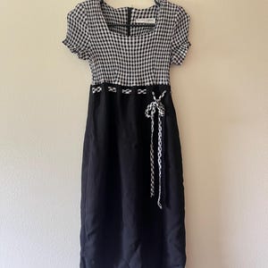 May include: A midi-length dress featuring a black and white gingham bodice with short sleeves and a square neckline. The solid black skirt is accented by a gingham waist trim and a matching tie detail. A label inside reads 100% THINGS.