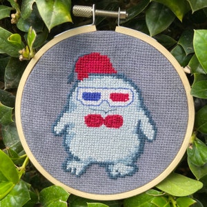 May include: A gray felt cross-stitch embroidery hoop with a white, gray, and red cartoon character wearing 3D glasses and a red hat. The character is wearing a red bow tie.