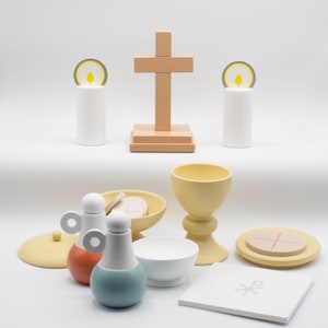 May include: A wooden cross with a light brown base, two white candles with yellow flames, a chalice, a paten, a ciborium, two cruets, and a white communion wafer. The items are arranged on a white surface.