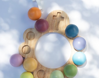 May include: A wooden rosary board with a central white circle and a cross-shaped cutout. It features colorful, round beads in shades of orange, purple, blue, green, yellow, and pink. A small white cross is also present.