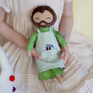 May include: A handcrafted doll with a smiling face, brown hair, and a beard. The doll is dressed in a green shirt and apron, with a pocket holding a hammer and ruler. The doll is held by a person wearing a cream-coloured dress.