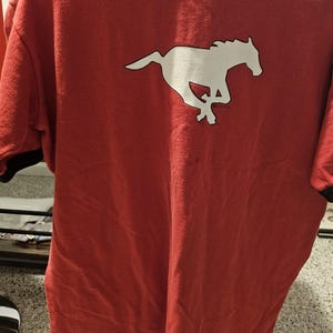 May include: Red t-shirt with a white running horse graphic. The shirt has a black trim around the collar and sleeves. The horse graphic is a silhouette of a galloping mustang.