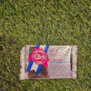 Puede incluir: Un paquete de aluminio plateado de tarjetas de colección de deportes 1991 Classic Draft Picks. El paquete presenta un logotipo rojo, blanco y azul con el texto "Classic Picks" y "1991 Draft". El paquete incluye 12 tarjetas de selección de draft de béisbol, baloncesto y fútbol, ​​más 10 tarjetas de bonificación especiales limitadas, incluido un set de 5 tarjetas Rocket Ismail.