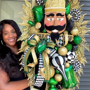 May include: A decorative nutcracker wreath featuring a central nutcracker figure with a green hat, black mustache, and gold accents. The wreath is adorned with gold fern leaves, green and gold ornaments, and black and white checkered ribbons.