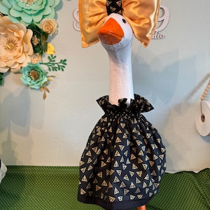May include: A decorative goose figurine with a white body, orange beak and feet, and a black dress with gold triangle patterns. It wears a large gold bow and a black collar. The goose is standing on a green base.