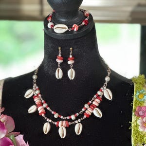 May include: A jewellery set consisting of a double-strand necklace, a bracelet, and drop earrings. Each piece features white cowrie shells, small red coral-like beads, and white pearls strung on a metallic chain with a lobster claw clasp.