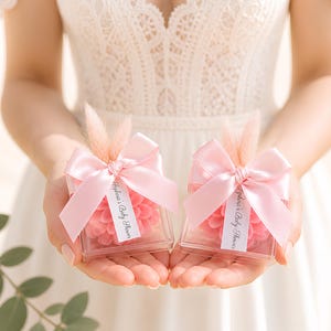 Puede incluir: Dos cajas cuadradas transparentes con arreglos florales rosas y lazos rosas. Cada caja tiene una pequeña etiqueta que dice "Sophia's Baby Shower". Las cajas se sostienen frente a un vestido blanco.
