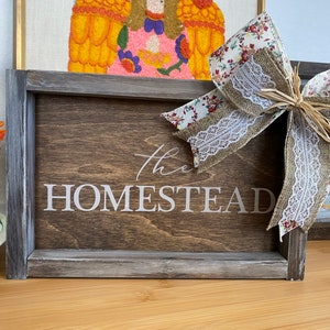 May include: A rustic wooden sign with a distressed gray frame reads "The Homestead" in white lettering. A large burlap and floral bow with white lace accents is tied to the sign.