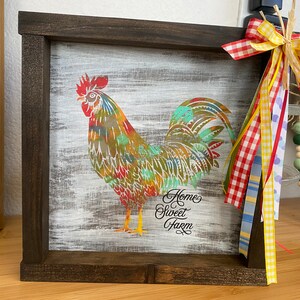 May include: A framed wooden sign with a colorful rooster illustration and the text "Home Sweet Farm". The sign is decorated with a red and white checkered ribbon, a yellow ribbon, and a light blue ribbon.