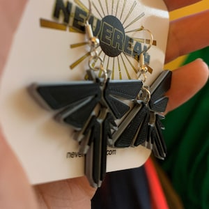 May include: A pair of black 3D printed earrings shaped like the Firefly logo from the video game The Last of Us Part II. The earrings are hanging from silver hooks and are on a white card with the text "NeverReal.com".