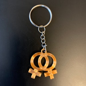 May include: A gold-colored keychain with two interlocking female symbols. The keychain is attached to a silver key ring and chain.