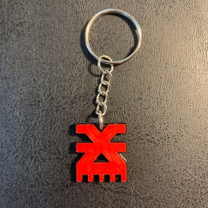 May include: A red keychain with a stylized symbol, resembling a geometric design, attached to a silver chain and ring.