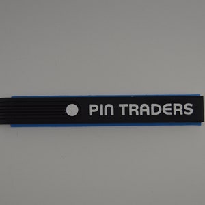 May include: A rectangular sign with the text "PIN TRADERS" in white letters. The sign has a black top layer, a blue middle layer, and a white dot on the left side. The left side of the sign has a series of vertical lines.