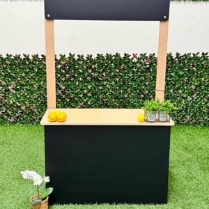 May include: A black and tan wooden lemonade stand with a black sign. The stand has a light-colored countertop with lemons and a small potted plant. An orchid in a woven basket sits in front of the stand.