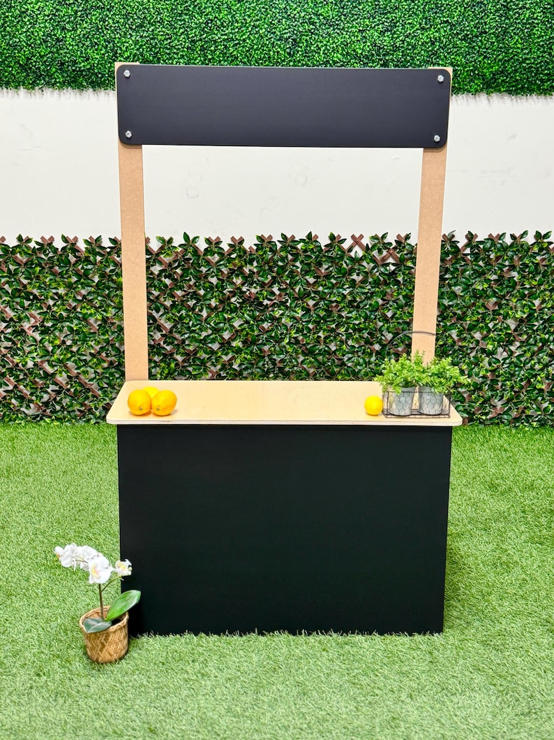 May include: A black and tan wooden lemonade stand with a black sign. The stand has a light-colored countertop with lemons and a small potted plant. An orchid in a woven basket sits in front of the stand.