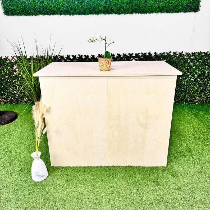 Pu&ograve; includere: Un bar in legno di colore chiaro con un piano piatto e lati angolati. Una piccola orchidea in vaso &egrave; appoggiata sopra. Il bar &egrave; su una superficie verde, con un muro bianco e una siepe verde sullo sfondo.