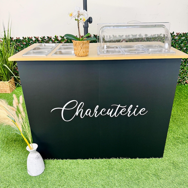 Pu&ograve; includere: Una stazione di servizio per salumi nera con un piano in legno chiaro. La stazione ha contenitori per alimenti in acciaio inossidabile e un coperchio in plastica trasparente. La parola "Charcuterie" &egrave; scritta in bianco sul davanti. Un'orchidea in vaso &egrave; in cima.