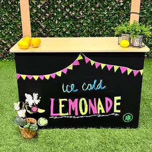 May include: A black lemonade stand with a wooden top and a green grass background. The stand has the words "ice cold LEMONADE" in colorful lettering. Yellow and pink pennant flags decorate the top. Lemons and plants are on the stand.