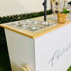 May include: A white serving cart with a light wood-effect top and stainless steel containers. The cart features the word "Charcuterie" in script. A small potted plant sits on the top surface.