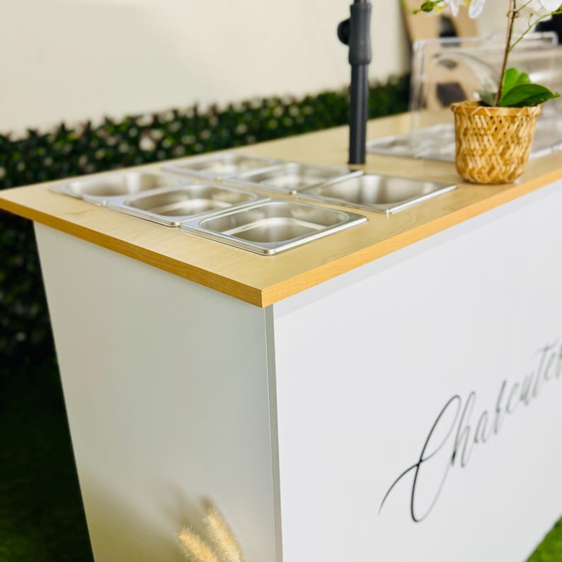 May include: A white serving cart with a light wood-effect top and stainless steel containers. The cart features the word "Charcuterie" in script. A small potted plant sits on the top surface.