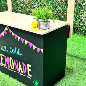 May include: A black lemonade stand with a light wood countertop. The stand has colorful pennant flags and the words "ice cold LEMONADE" written on the front. A small potted plant, a lemon, and a wire basket are on the countertop.