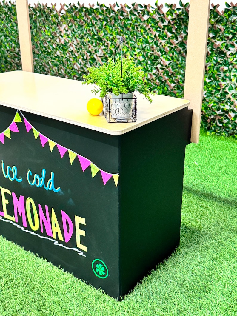 May include: A black lemonade stand with a light wood countertop. The stand has colorful pennant flags and the words "ice cold LEMONADE" written on the front. A small potted plant, a lemon, and a wire basket are on the countertop.